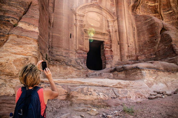 A person, holding a smartphone, photographs a carved stone facade with a large, dark entrance in an ancient archaeological site, surrounded by rocky, sunlit surfaces.