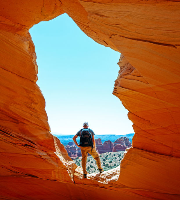 A person with a backpack stands on a rock ledge, framed by an orange rock formation, gazing at distant rugged sandstone landscapes under a clear blue sky.