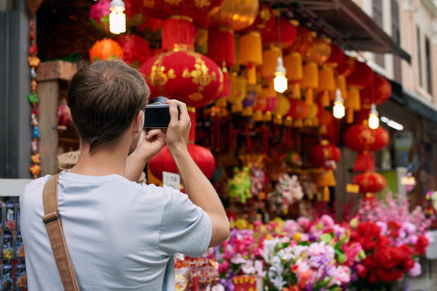 Person photographing vibrant red and yellow lanterns hanging outside a shop. Surrounding colorful artificial flowers and festive decorations enhance the lively market atmosphere.