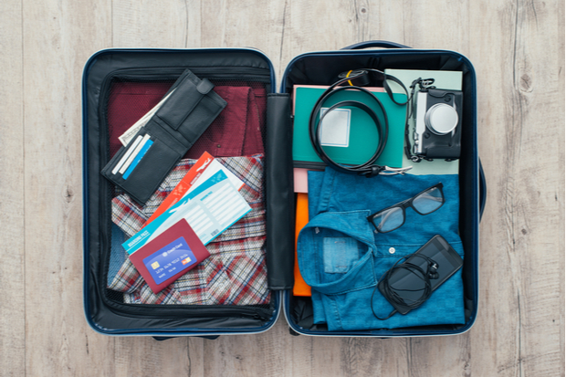 Suitcase packed with folded clothes, travel documents, a wallet, glasses, camera, and charging cables, rests open on a wooden floor, ready for travel.