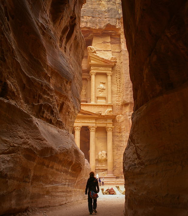 A person walks through a narrow sandstone gorge towards an ancient, ornately carved building facade, with columns and sculptures, in a sunlit, historic archaeological site.