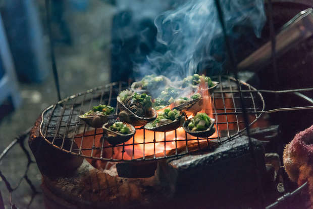 Shellfish are grilling on a metal grate over an open flame. Smoke rises, surrounding them. Finely chopped green herbs garnish the shellfish, suggesting a casual outdoor cooking setting.