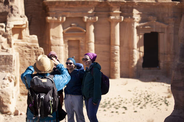 A person photographs three friends standing together, smiling, wearing casual outdoor clothing and sunglasses, against a backdrop of ancient stone architecture in a sunlit desert setting.