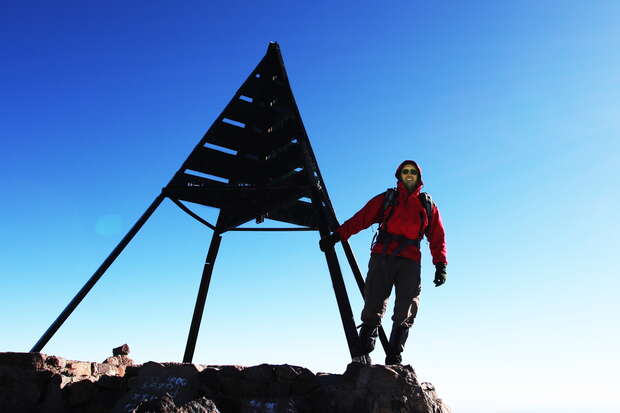 A person wearing a red jacket stands on a rocky summit, touching a triangular metal marker. The sky is clear and blue, creating a striking backdrop.