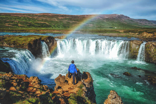 A person in a blue jacket stands on a rocky cliff, observing a waterfall with a vibrant rainbow. Lush greenery and mountains create a scenic backdrop under a partly cloudy sky.