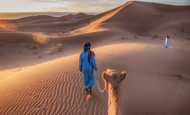 A person in blue robes leads a camel across rippled sand dunes at sunset. Another person is visible in the distance, with expansive desert surrounding them.