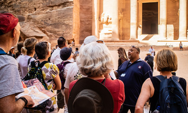 A tour group listens attentively to a guide in an ancient city, surrounded by towering sandstone cliffs and elaborate carved stone structures, under natural sunlight.