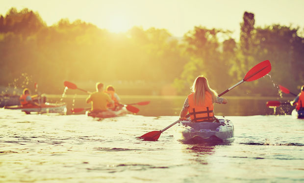 Kayakers paddle across a sunlit lake, surrounded by trees. They wear life jackets while maneuvering their boats in the tranquil water, enjoying the serene outdoor setting.