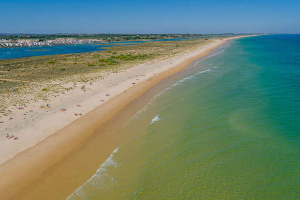 A long, sandy beach stretches alongside calm, blue-green ocean waves, with scattered people sunbathing. In the background, a river leads to a distant town under a clear blue sky.