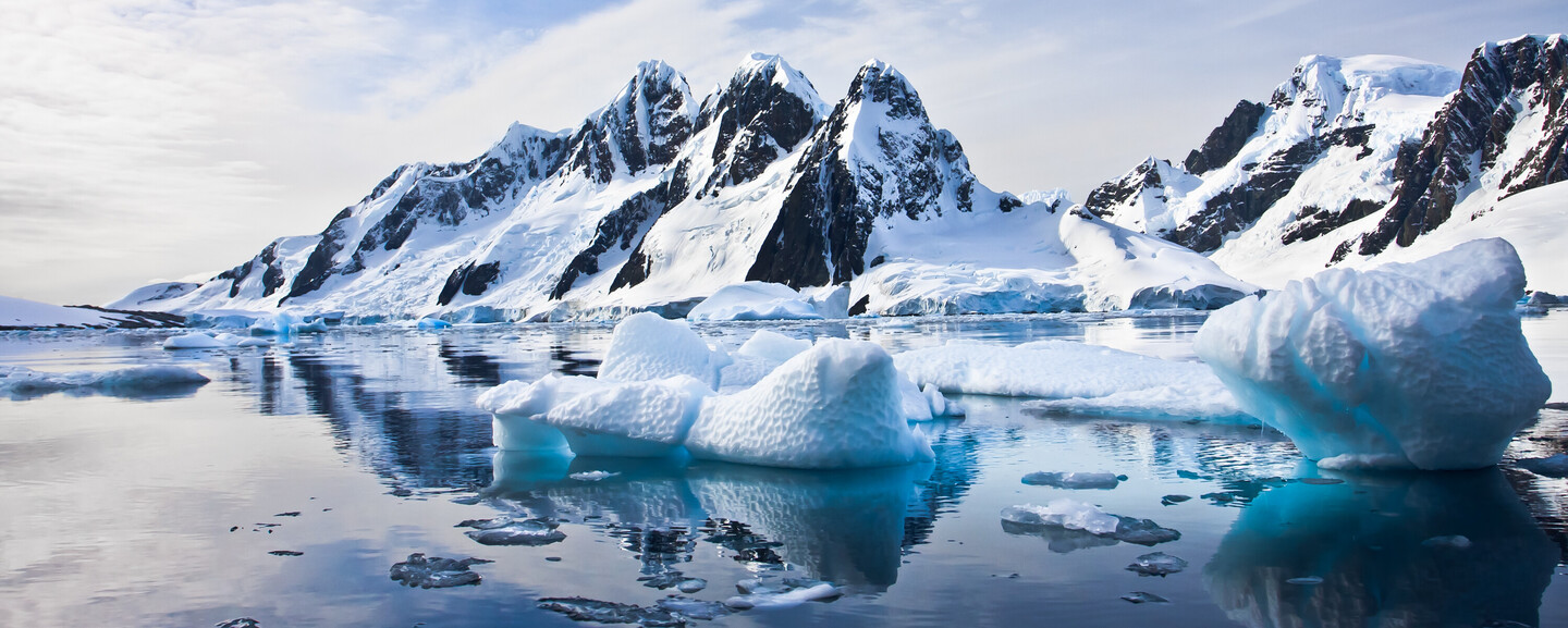 Icebergs float in calm, reflective water alongside jagged, snow-covered mountains under a partly cloudy sky, creating a serene, icy landscape scene.