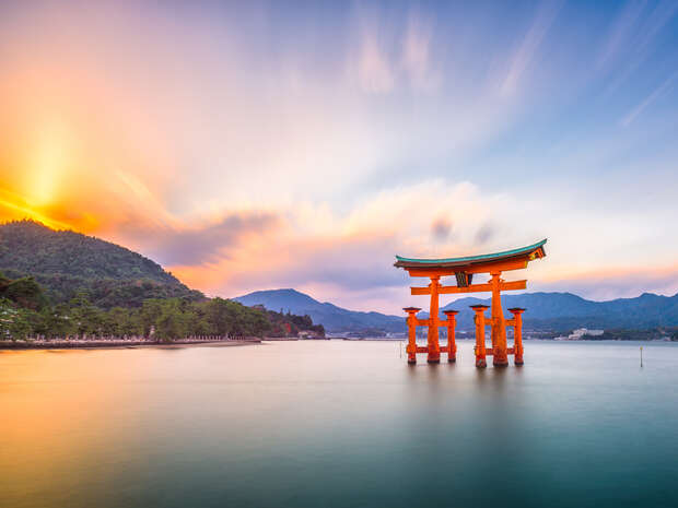 A large orange torii gate stands in calm water, with a vibrant sunset sky above and forested mountains in the distance, creating a serene and picturesque landscape.
