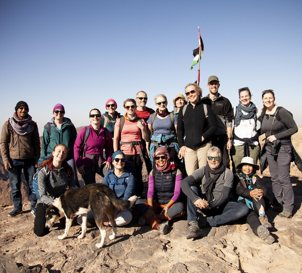 A diverse group of people, standing and smiling, pose on a rocky plateau. A dog is present, and a flag waves in the background under a clear blue sky.