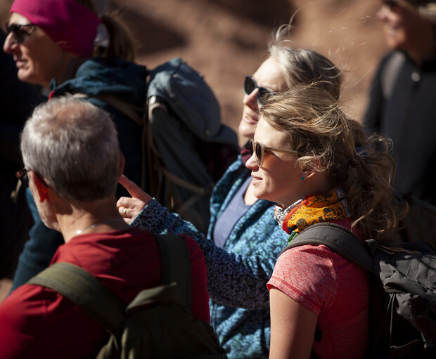 A group of people wearing backpacks and sunglasses walk outdoors. One person gestures while others look around, suggesting a hiking activity in a sunny, natural setting.
