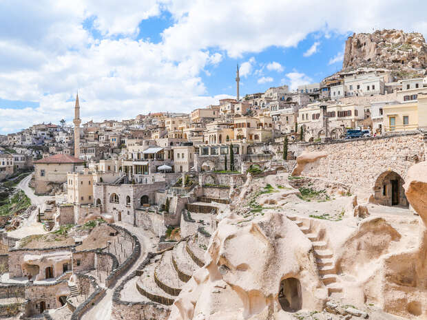 Rocky dwellings sit on a hillside, with a labyrinth of stone buildings and a distant mosque rising above. The scene is under a partly cloudy sky in a historic town.