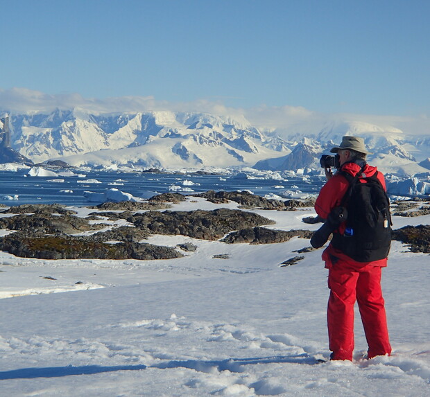 A person in red winter gear photographs snow-covered mountains and icy waters, surrounded by a vast, frozen landscape under a clear blue sky.