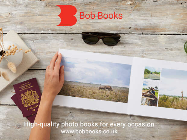 A hand flips through a photo book featuring safari images on a wooden table with sunglasses, passports, and dried flowers. Text reads: "Bob Books. High-quality photo books for every occasion. www.bobbooks.co.uk."