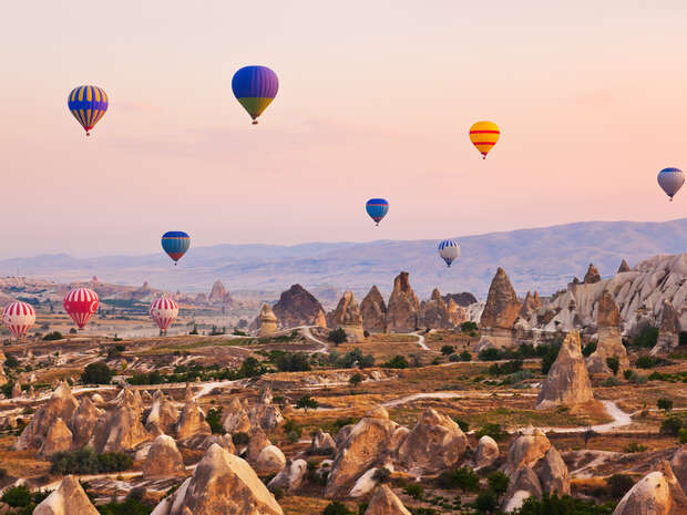 Hot air balloons float above a rocky landscape with unique, tall formations; the sky is softly lit, suggesting either sunrise or sunset, providing a serene and picturesque backdrop.
