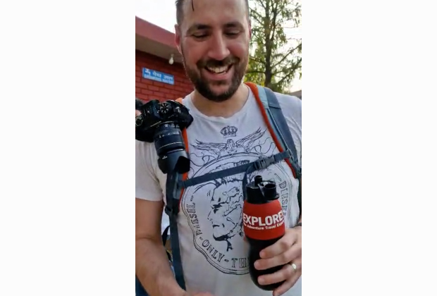 A person smiles while holding a bottle labeled "EXPLORE." A camera hangs from their shoulder, and they're wearing a printed shirt. A brick building and trees are in the background.