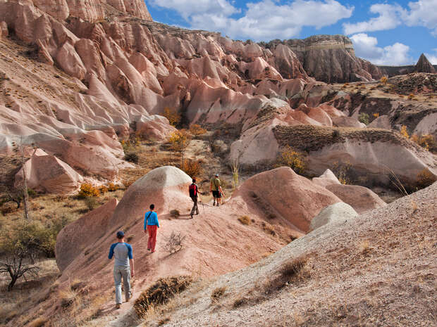 Hikers walk on a dusty trail among unique, pink-hued rock formations and sparse vegetation under a partly cloudy sky.