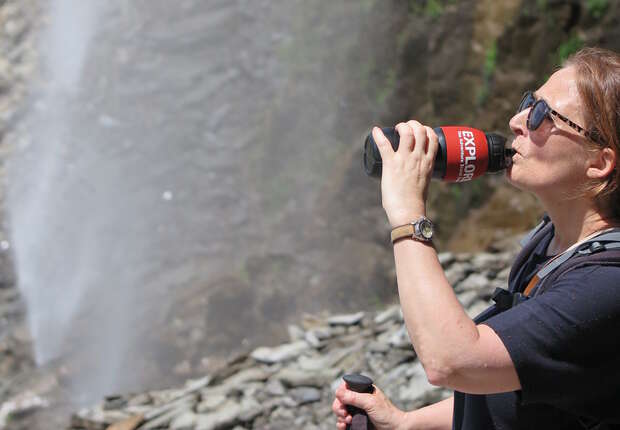 A person drinks from a bottle labeled "EXPLORE" near a rocky waterfall, wearing sunglasses and a backpack, and holding a walking stick.