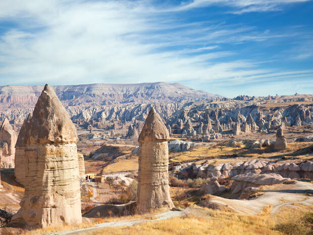 Conical rock formations stand prominently, rising from a dry, grassy landscape with scattered vegetation, against a backdrop of distant hills and a bright blue sky.