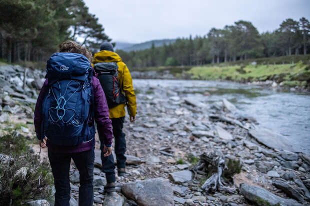 Two people with backpacks walk along a rocky riverbank, surrounded by trees and distant hills. One has a visible blue backpack with a design on it.
