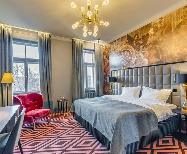 A modern bedroom features a large, neatly made bed with gray bedding. A striking chandelier hangs overhead. The room includes a patterned carpet, artistic wallpaper, windows with curtains, and a red chair.