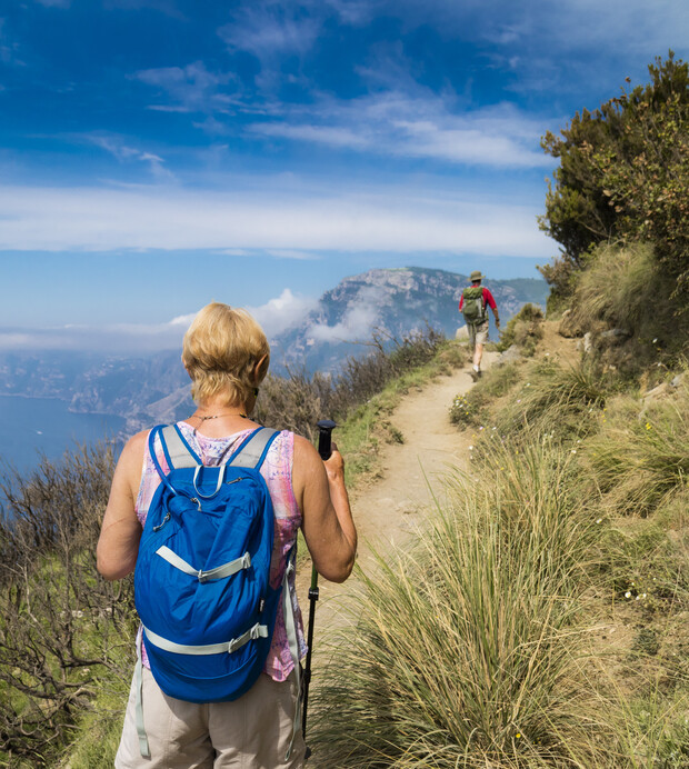 A hiker with a blue backpack and hiking pole walks on a narrow dirt path. The path is surrounded by tall grass, with mountains and blue sky in the background.
