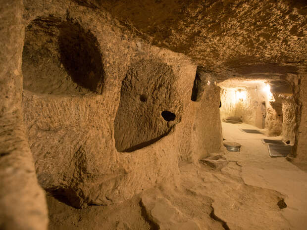 Rock-hewn tunnels extend through an underground passage, lit softly by artificial lights, creating a network of interconnected chambers with rough, stone walls in a dimly lit environment.