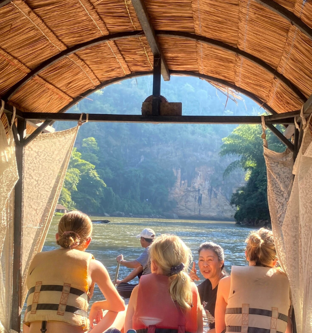People in life jackets sit on a raft under a thatched canopy, floating down a river surrounded by lush, green cliffs. Bright sunlight illuminates the scene.