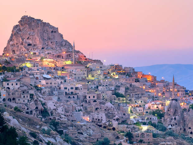 Rocky hillside town with ancient stone buildings illuminated by scattered lights, creating a warm, picturesque glow. The pink and blue sky hints at twilight, adding a serene atmosphere.