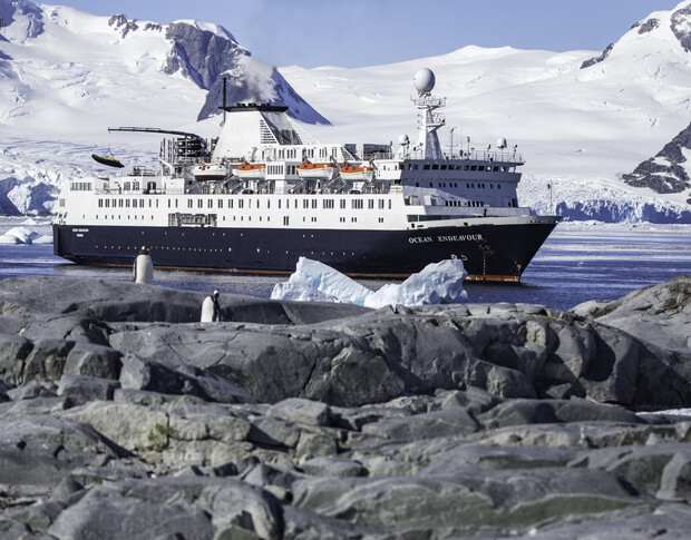 Ship "Ocean Endeavour" navigates icy waters, passing icebergs. Mountainous, snow-covered terrain forms the backdrop. In the foreground, rocks and two penguins are visible, enhancing the polar scene.
