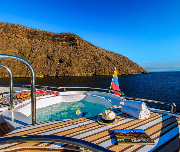 A jacuzzi on a yacht deck contains water and a cocktail with a lemon slice. Nearby are rolled towels, a hat, and a book. A Colombian flag waves against a rocky island backdrop under clear skies.