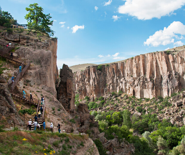 People walk down a wooden staircase built into a rocky cliffside with a lush valley below, under a bright blue sky with scattered clouds.