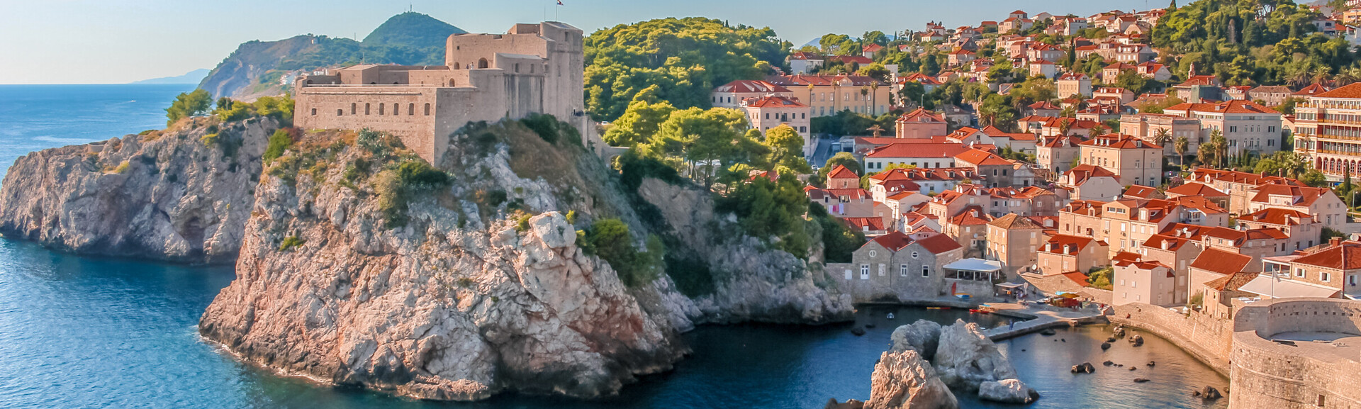 Fortress sits on a rocky cliff, overlooking the sea. Red-roofed buildings fill the hillside, surrounded by green trees under a clear blue sky. A small harbor is visible below.