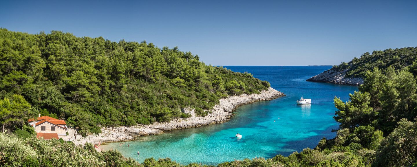 A boat floats on clear blue water in a secluded bay, surrounded by lush green trees and a rocky shoreline. A small house with a red roof sits on the left.