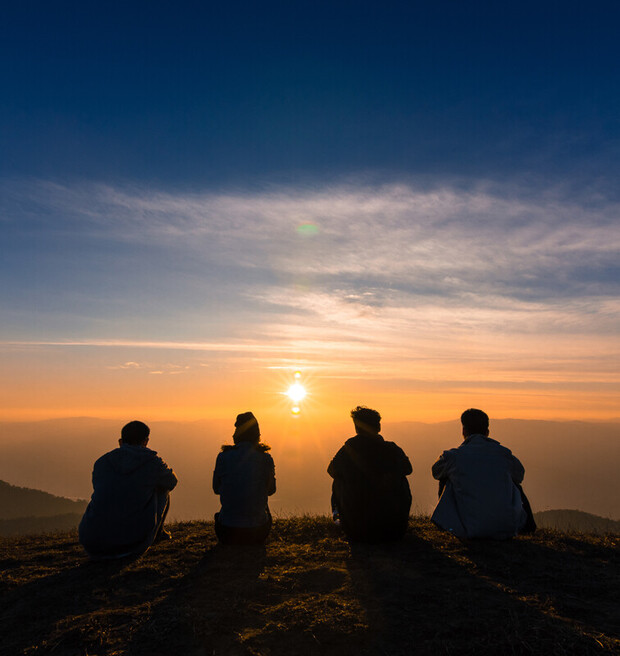 Four people sit on a hilltop, watching the sun set over distant mountains under a colorful sky with orange and blue hues.