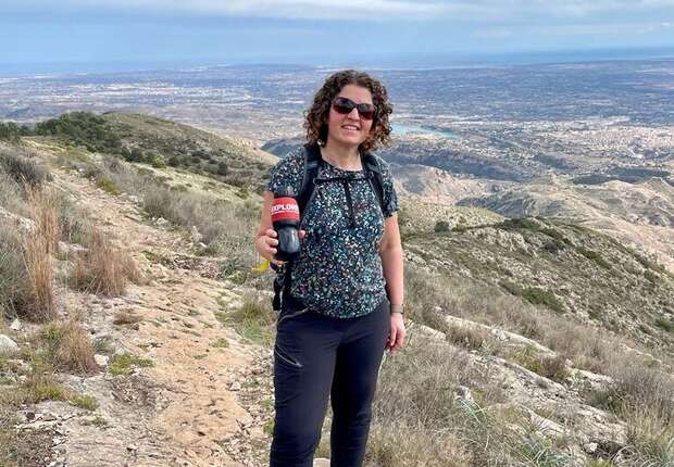 A woman stands on a mountain trail holding a red bottle labeled “Explora,” with a panoramic view of a distant city under cloudy skies in the background.