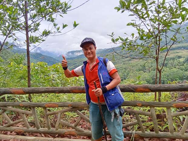A person stands, giving a thumbs-up and holding a walking stick, wearing casual clothes and a cap, in a lush, mountainous landscape with trees and a wooden fence.