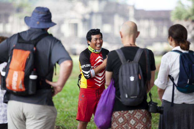 A smiling person wearing a colorful sports outfit interacts with a group of people in casual attire. They stand outdoors on a grassy area, with an ancient stone structure blurred in the background.