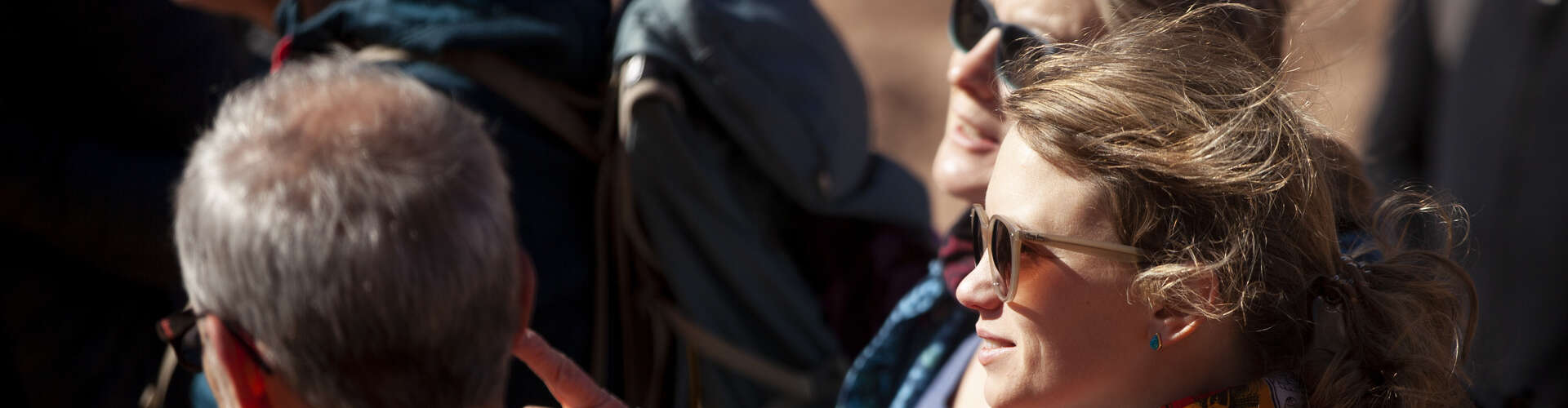 Several people, wearing sunglasses and casual clothing, look upwards in a sunny outdoor setting, possibly observing something in the sky or above them.