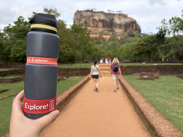 A hand holds a gray water bottle labeled "Explore!" while hikers walk along a path towards a large rock formation surrounded by greenery and trees, under a cloudy sky.
