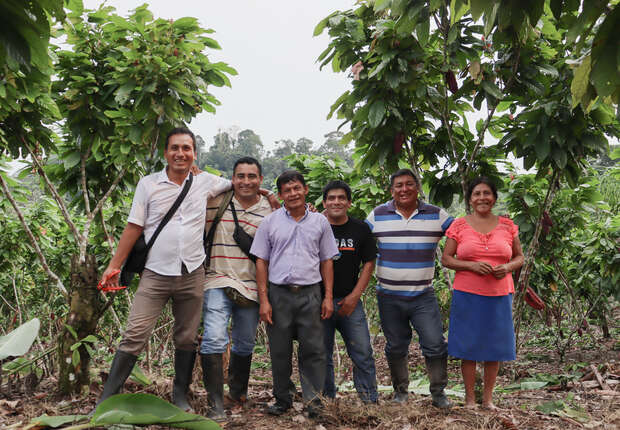 Seven people stand smiling among lush green trees, dressed casually, with backpacks and boots, suggesting they are in a tropical orchard or farm area.