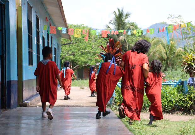 Children and an adult walk along a blue building's corridor, dressed in red garments. Colorful flags hang above. Lush greenery and hills are visible in the background.