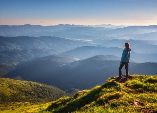 A person stands on a grassy mountain edge, gazing over a vast expanse of layered blue mountains under a clear sky, evoking a sense of tranquility and vastness.