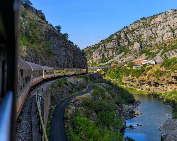 A train travels along a curving track beside a river, nestled between rocky cliffs. The landscape features lush greenery and a clear blue sky, with buildings visible in the distance.