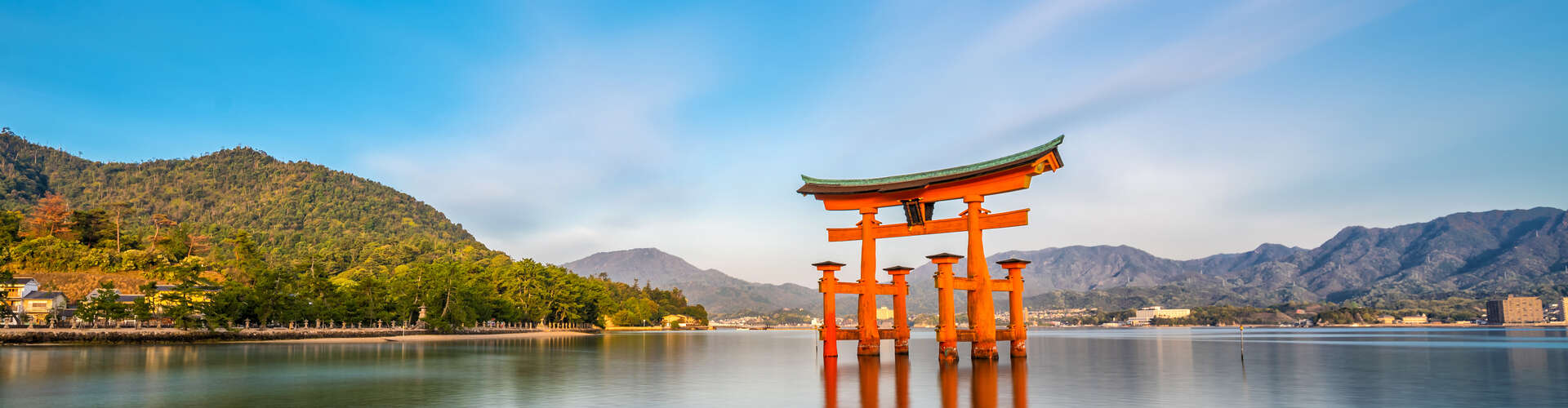A large, red torii gate stands in calm, reflective water, surrounded by mountainous, green landscapes and a clear blue sky.