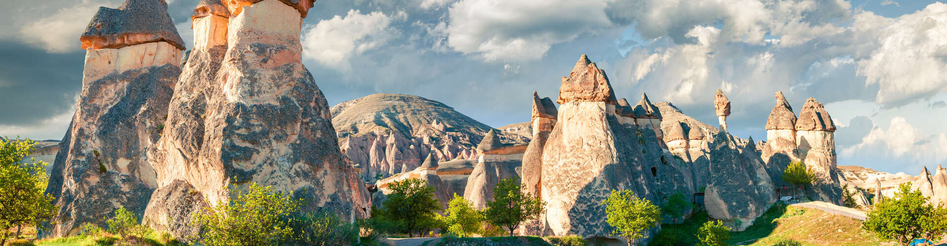 Rock formations tower, resembling fairy chimneys, under a partly cloudy sky. Scattered greenery grows around the formations, set against distant rolling hills.