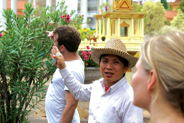 A person in a straw hat gestures while speaking to two other people in a garden setting with trees and a decorative yellow structure in the background.