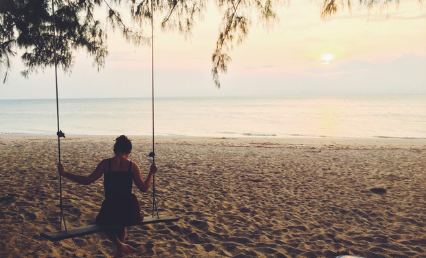 A person sits on a swing, gazing at the ocean during sunset on a sandy beach, with overhanging tree branches above.